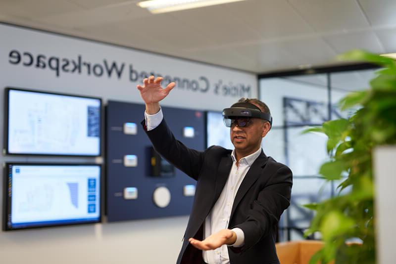 Man, wearing a black suit and white shirt, with a VR headset on his head and waving his arms. 'Mitie's Connected Workspace' is printed on the wall behind him